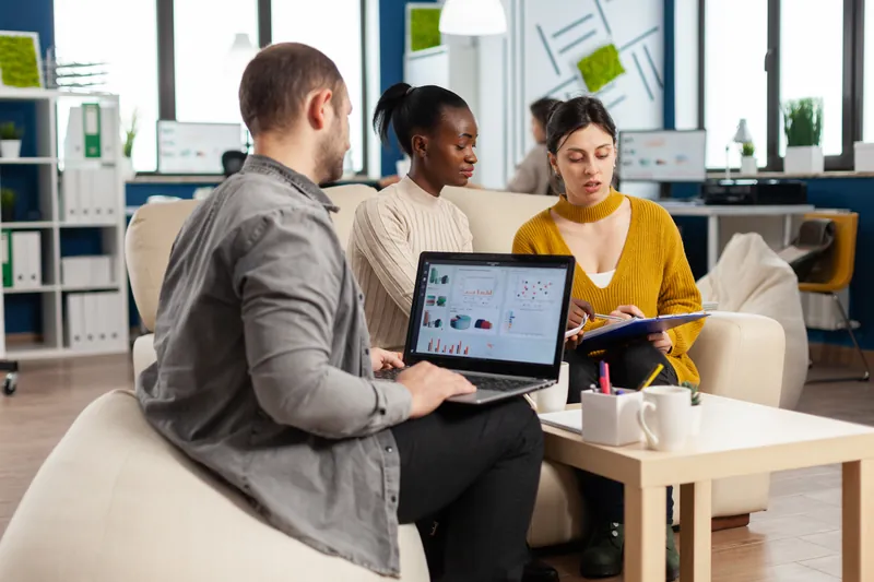 Three colleagues on an office sofa discussing charts on a laptop screen