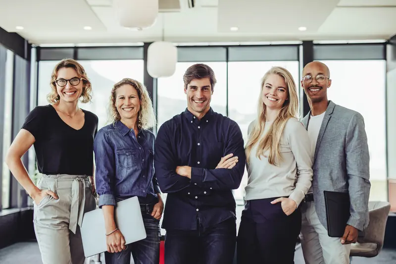 Five colleagues standing together in a bright office, two of them holding laptops
